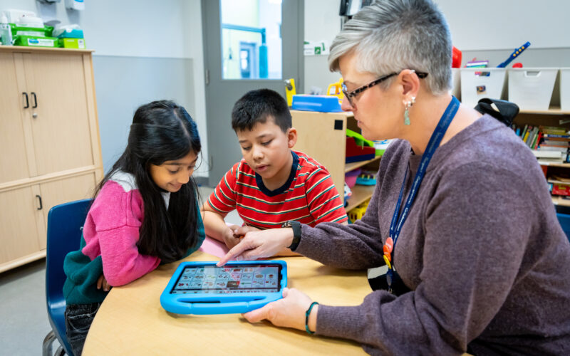 A young girl (left) with long dark hair and a young boy (middle) with a red striped shirt are sitting on a wooden table looking at an assistive communications device for children with autism. There are various images on the screen as a middle-aged woman (rights) points to one of the images.